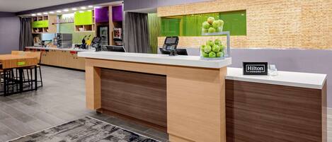 Zona de recepción de hotel con mostrador, quiosco digital y una exhibición de manzanas verdes.