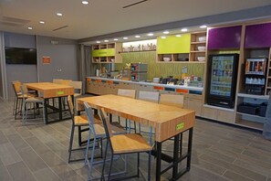 A restaurant with wooden tables and chairs, a counter with a coffee machine, and a refrigerator with beverages. - Home2 Suites by Hilton Salisbury (Salisbury)