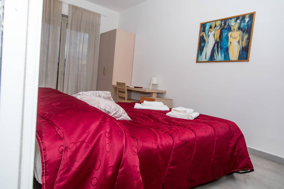 Un dormitorio con una colcha roja, un escritorio de madera, una silla, una lámpara blanca y un cuadro en la pared.