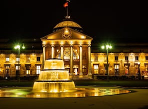 Um edifício imponente com cúpula e colunas, uma fonte em frente e uma bandeira no topo.