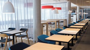Un espace de restauration moderne avec de longues tables et des chaises, de grandes fenêtres ornées de rideaux légers et des luminaires suspendus.