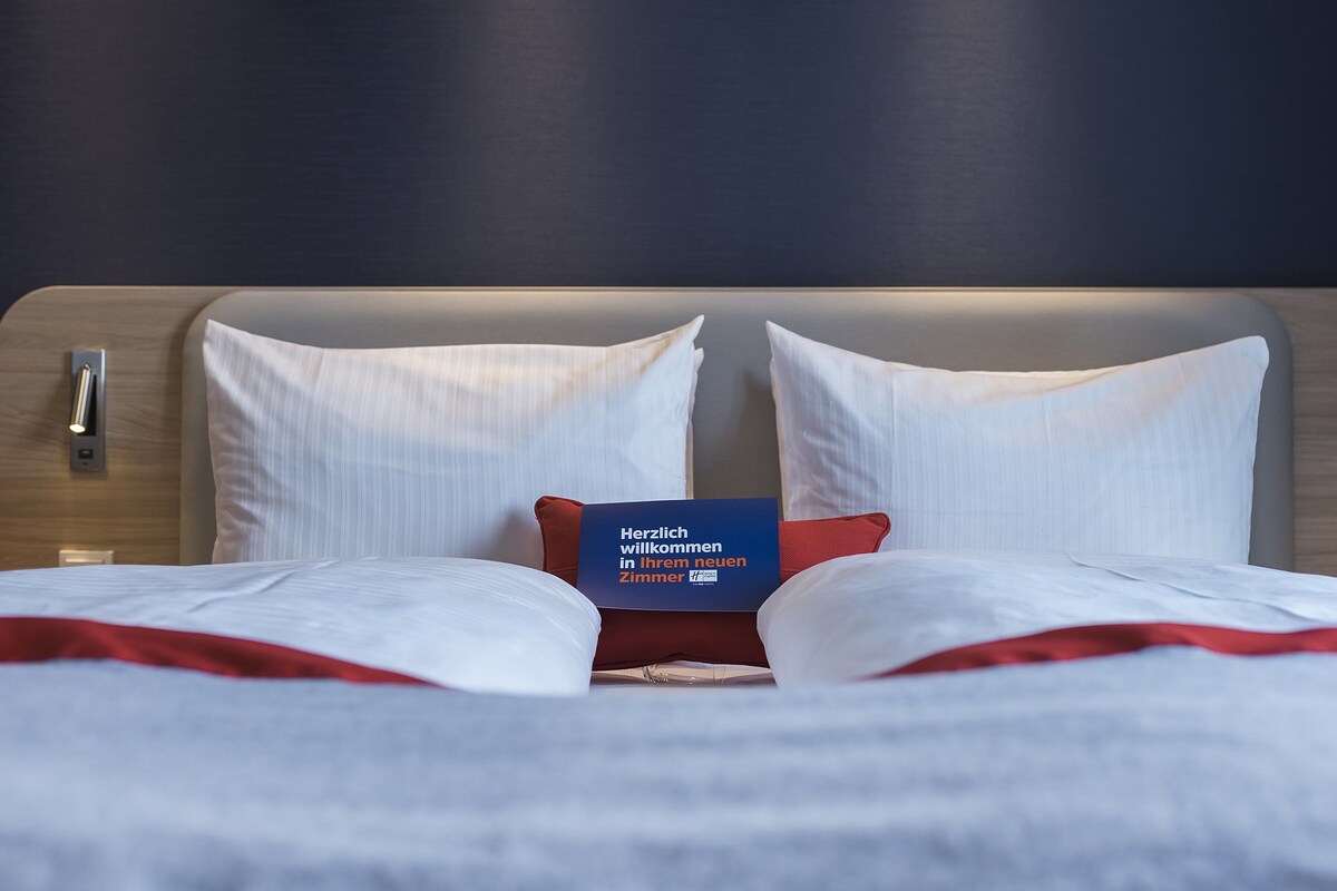 Een net opgemaakt bed met witte lakens en een rood kussen, een blauw welkomstbord en wandlampen.