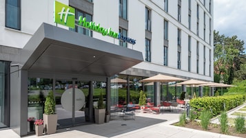 Entrada de un hotel con fachada de vidrio, zona de estar al aire libre y un letrero que dice 'Holiday Inn Express'.