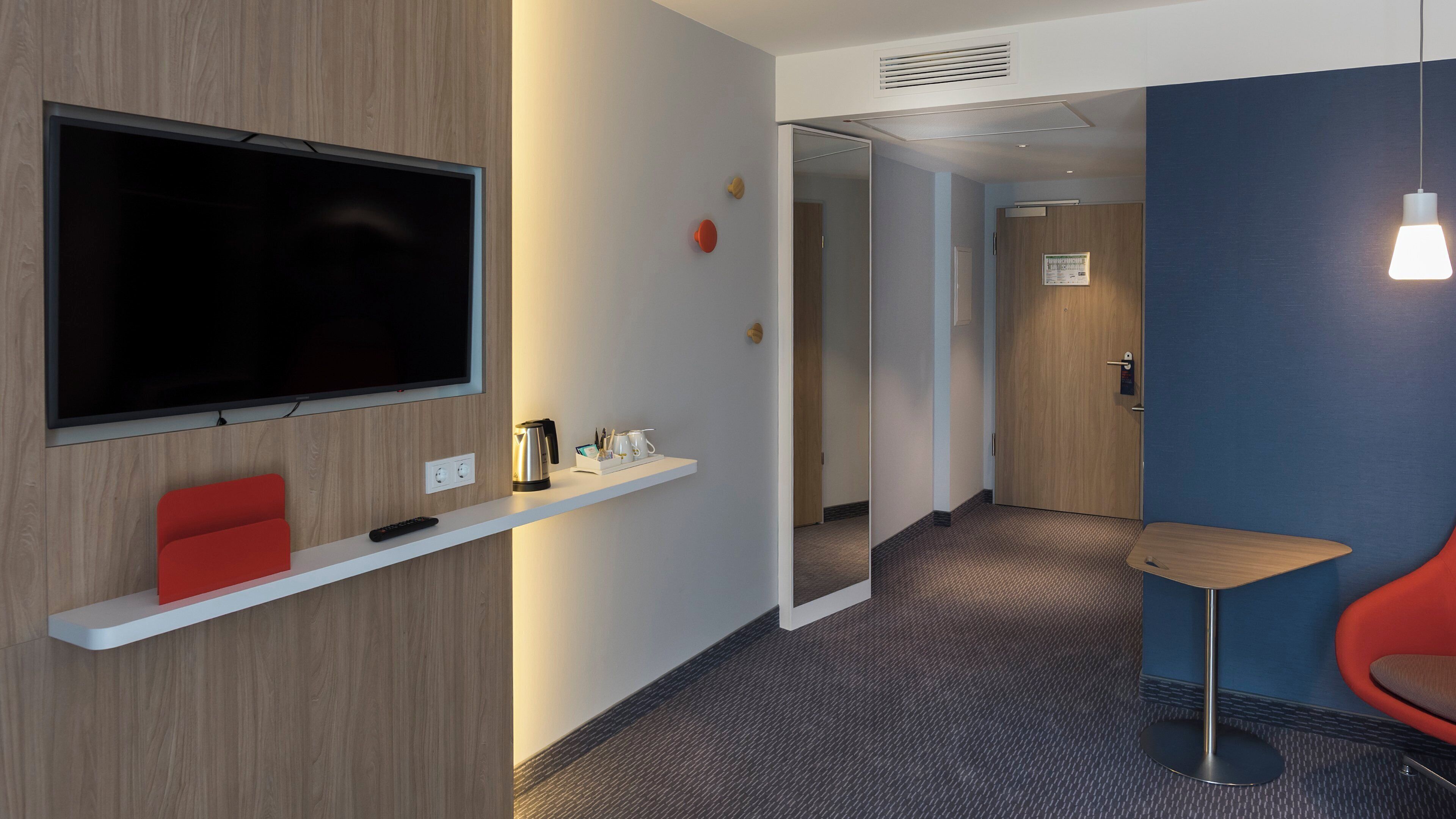 Une chambre d’hôtel équipée d’un téléviseur à écran plat, d’un fauteuil rouge, d’une petite table et d’une étagère sur laquelle se trouvent une bouilloire et des tasses.