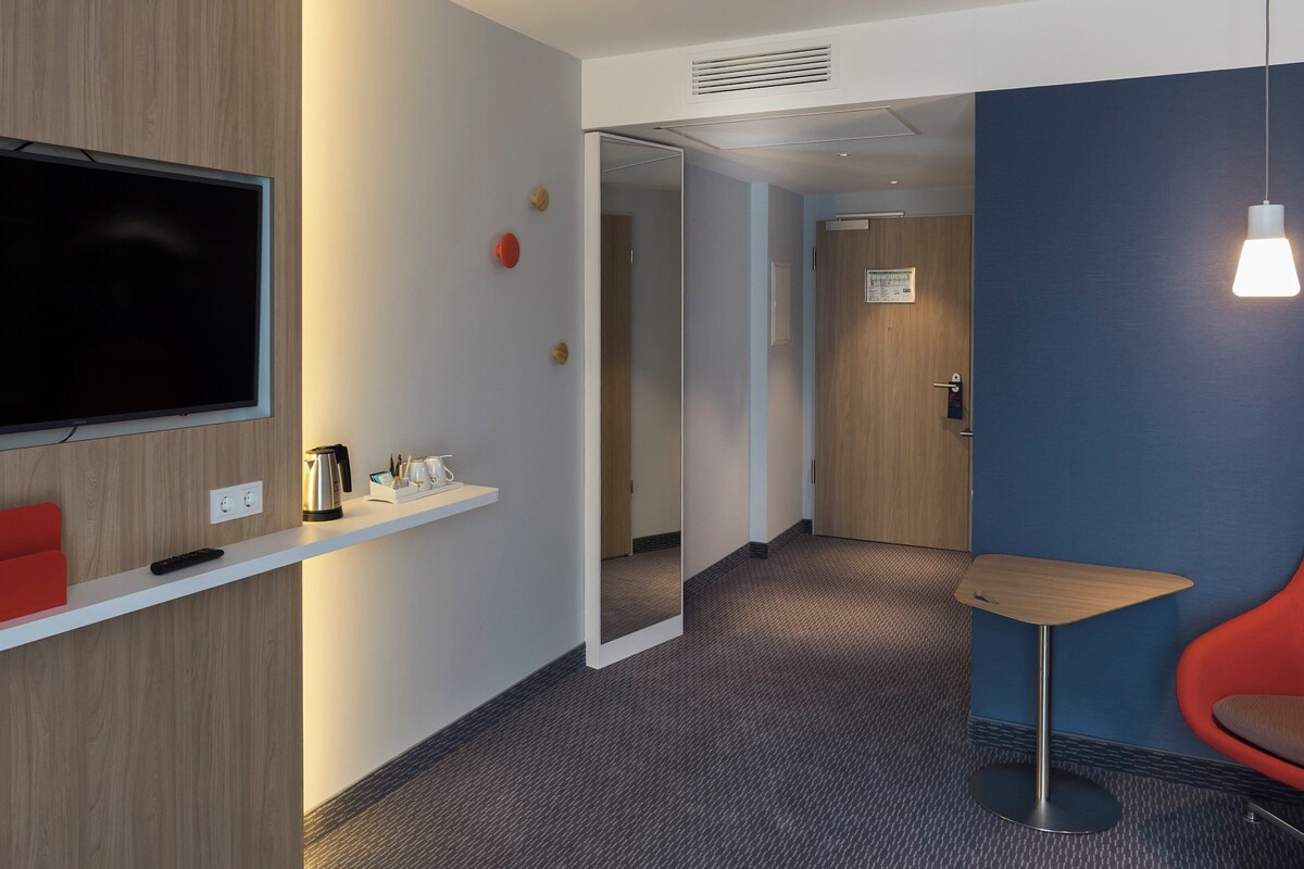 Een hotelkamer met een aan de muur bevestigde televisie, een kleine rode stoel, een witte plank met een waterkoker en kopjes, en een blauwe accentmuur.