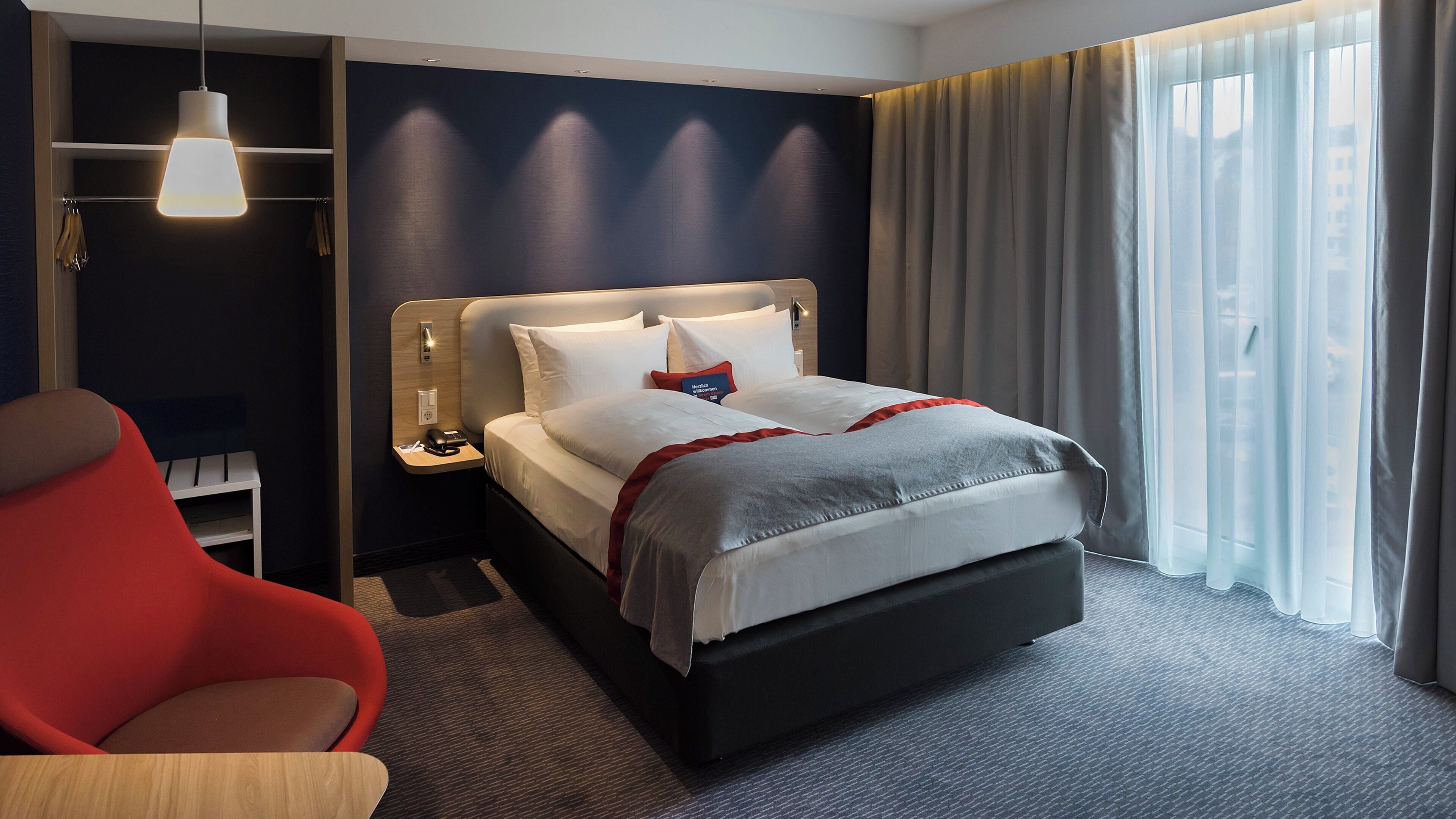 A hotel room with a bed, bedside table, lamp, chair, and a view of the outdoors through the window.