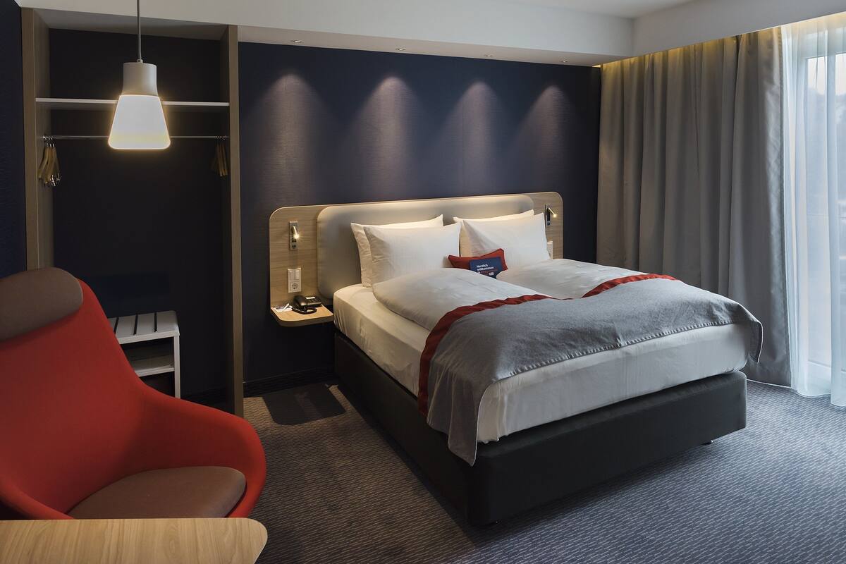 Habitación de hotel con cama, una silla roja, una mesita de noche con lámpara y ventana con cortinas.