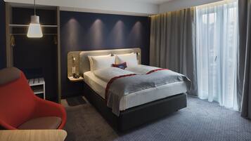 A hotel room with a bed, a red chair, a bedside table with a lamp, and a window with curtains.