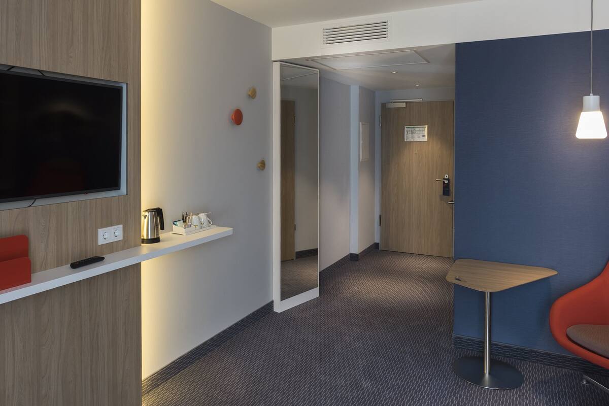 Una habitación de hotel con un televisor montado en la pared, una pequeña silla roja, un estante blanco con una tetera y tazas, y una pared de acento azul.