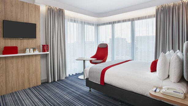 Une chambre d’hôtel avec un lit, une chaise, une petite table, un téléviseur, une étagère avec quelques objets et de grandes fenêtres.