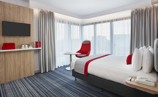 Quarto de hotel com uma cama, uma cadeira, uma mesinha, televisão, prateleira com objetos e janelas amplas.