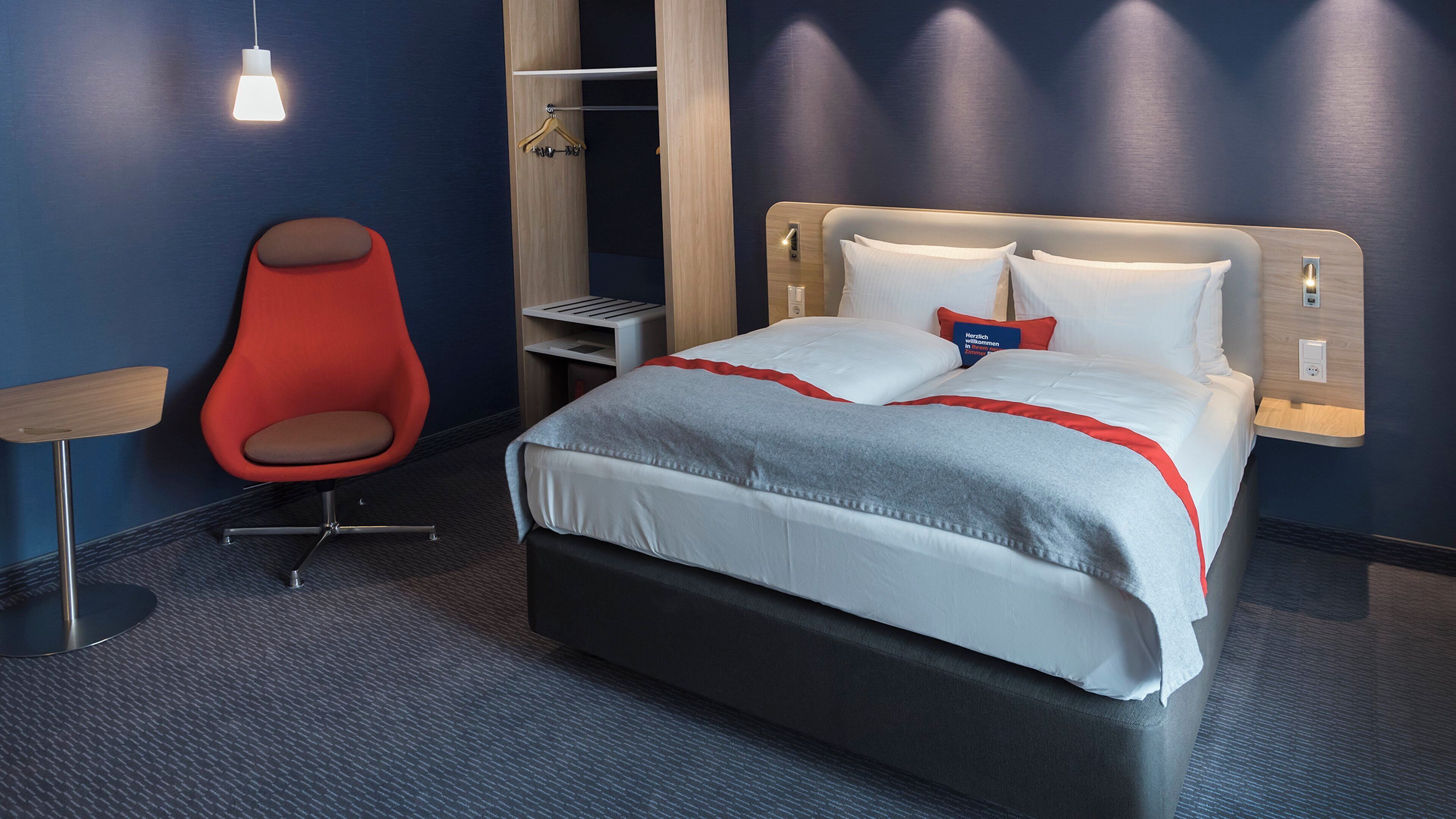 A hotel room with a bed, a red chair, a bedside table, and a wardrobe.