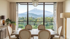 Un comedor con una mesa grande y cuatro sillas, una araña, una planta en maceta y vistas de una montaña a través de amplios ventanales.