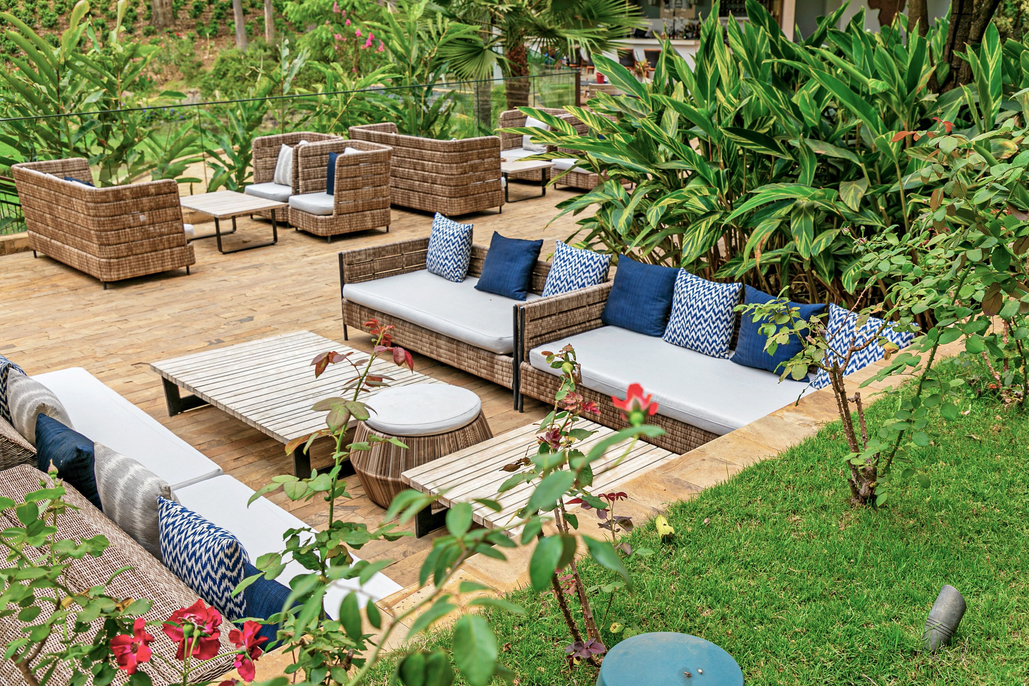 An outdoor patio area with wicker furniture, including sofas and a dining table, surrounded by lush greenery.