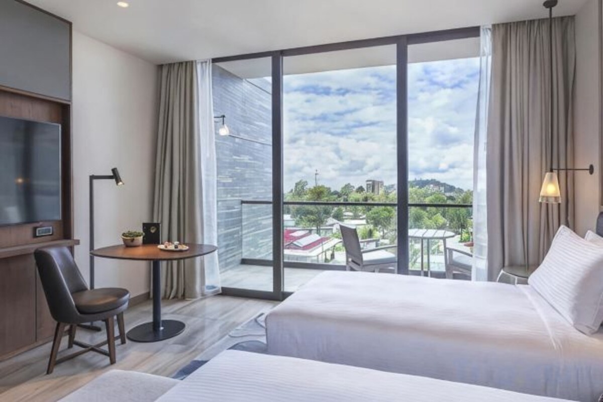 A hotel room with two beds, a TV, a small table with a chair, and a balcony with a view.