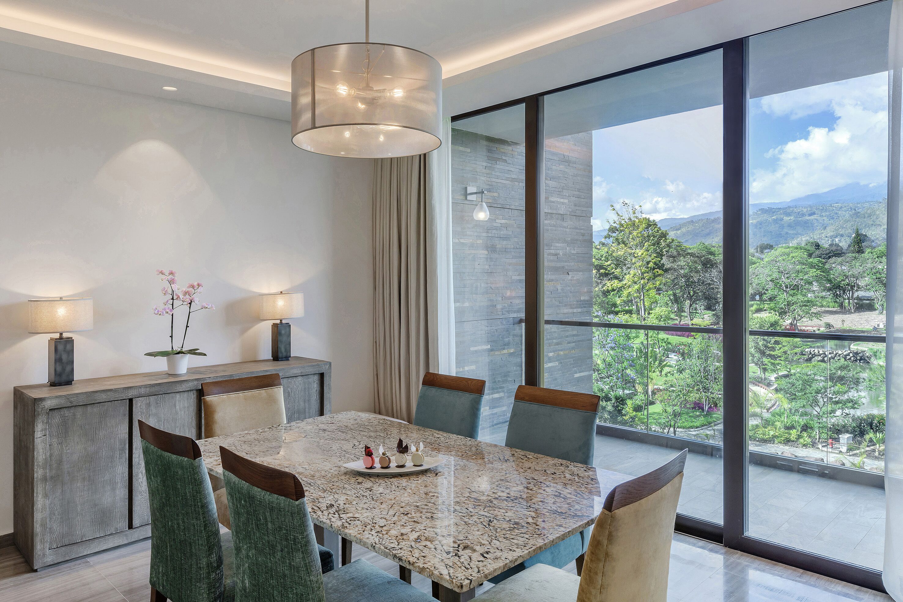 A modern dining area with a marble table, green upholstered chairs, and a view of a lush landscape through large windows.