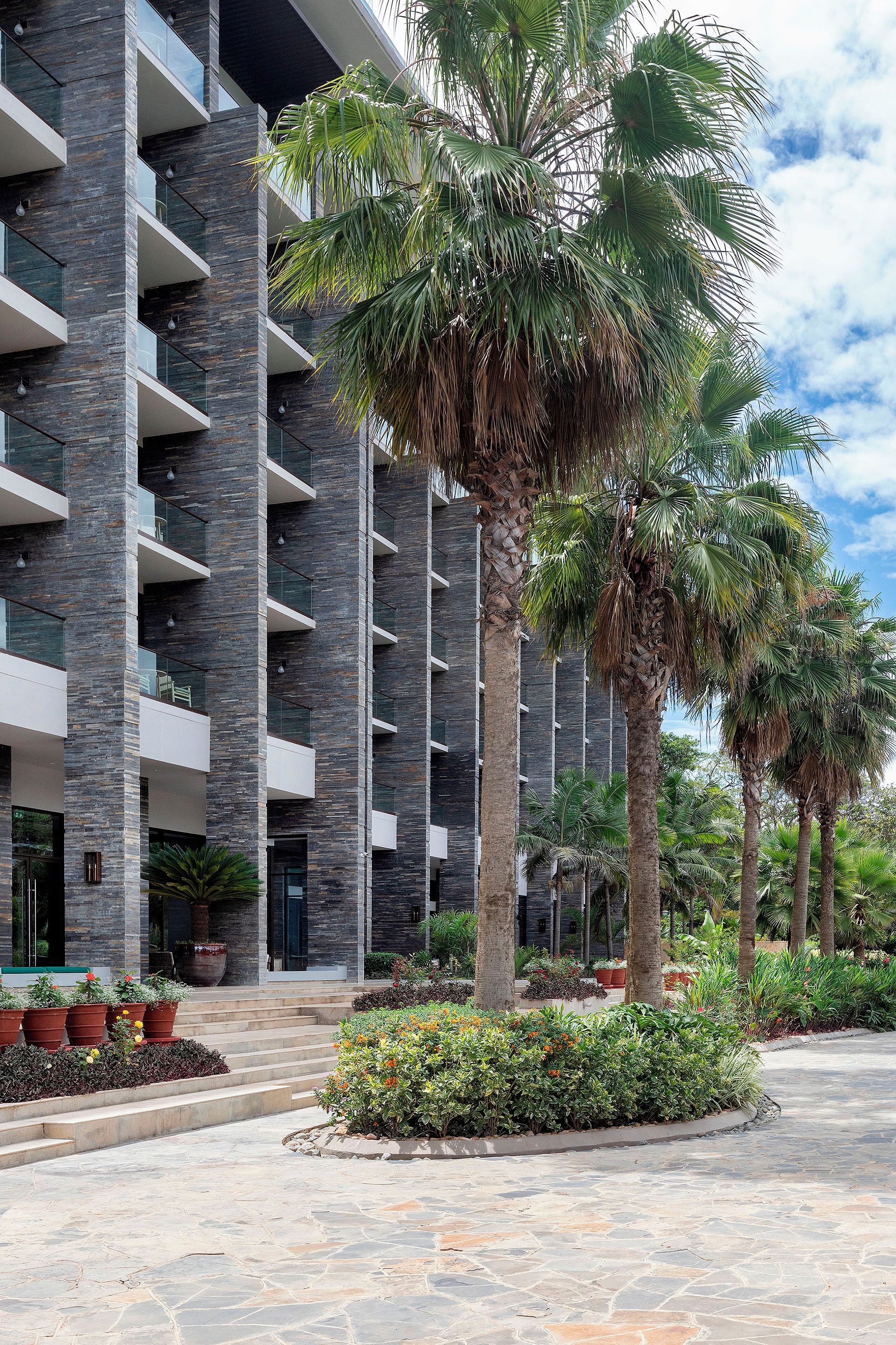 Un edificio de varios pisos con balcones y un patio ajardinado con palmeras y plantas en macetas.