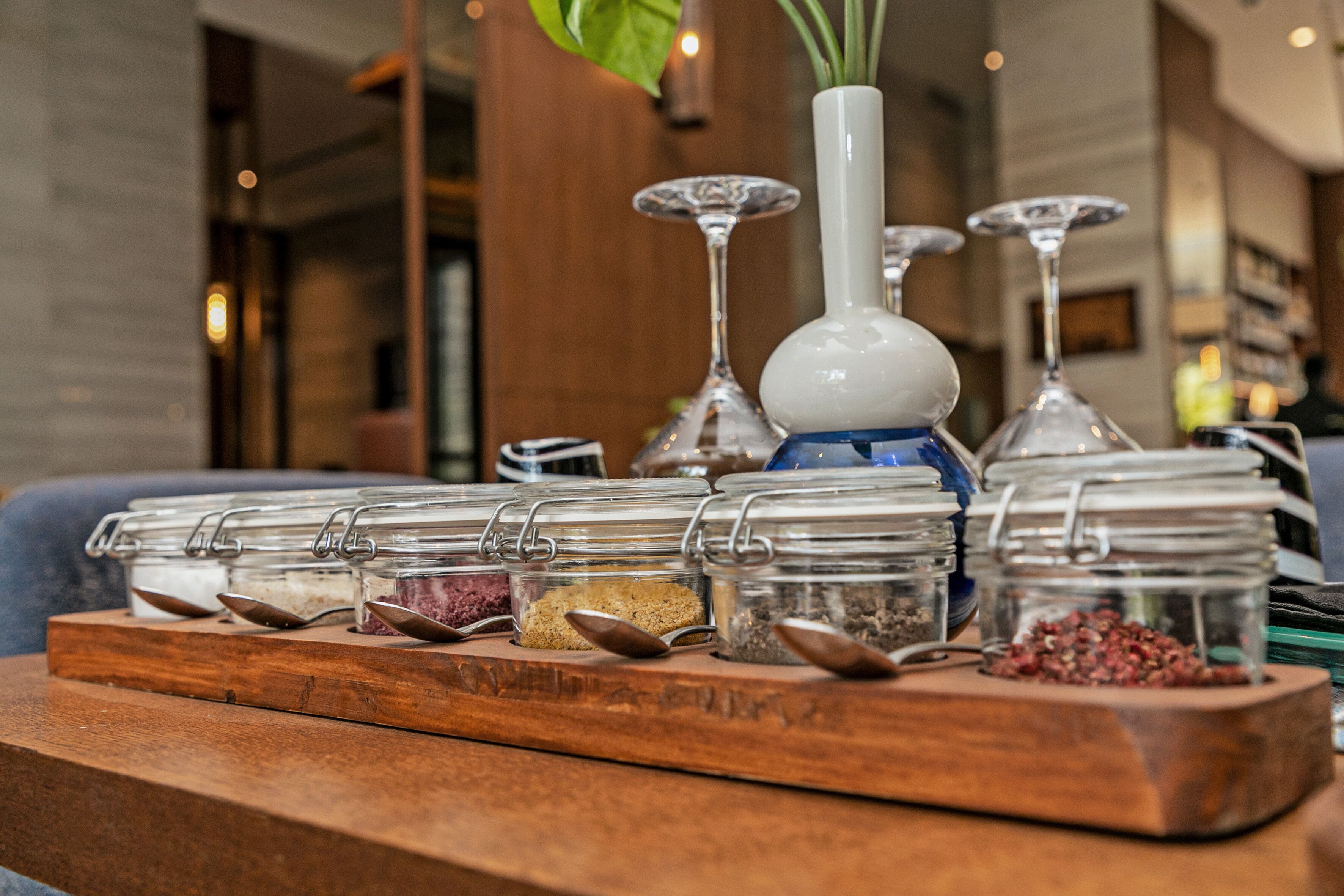 A wooden tray with multiple glass jars containing various food items, a white vase with a plant, and several wine glasses.