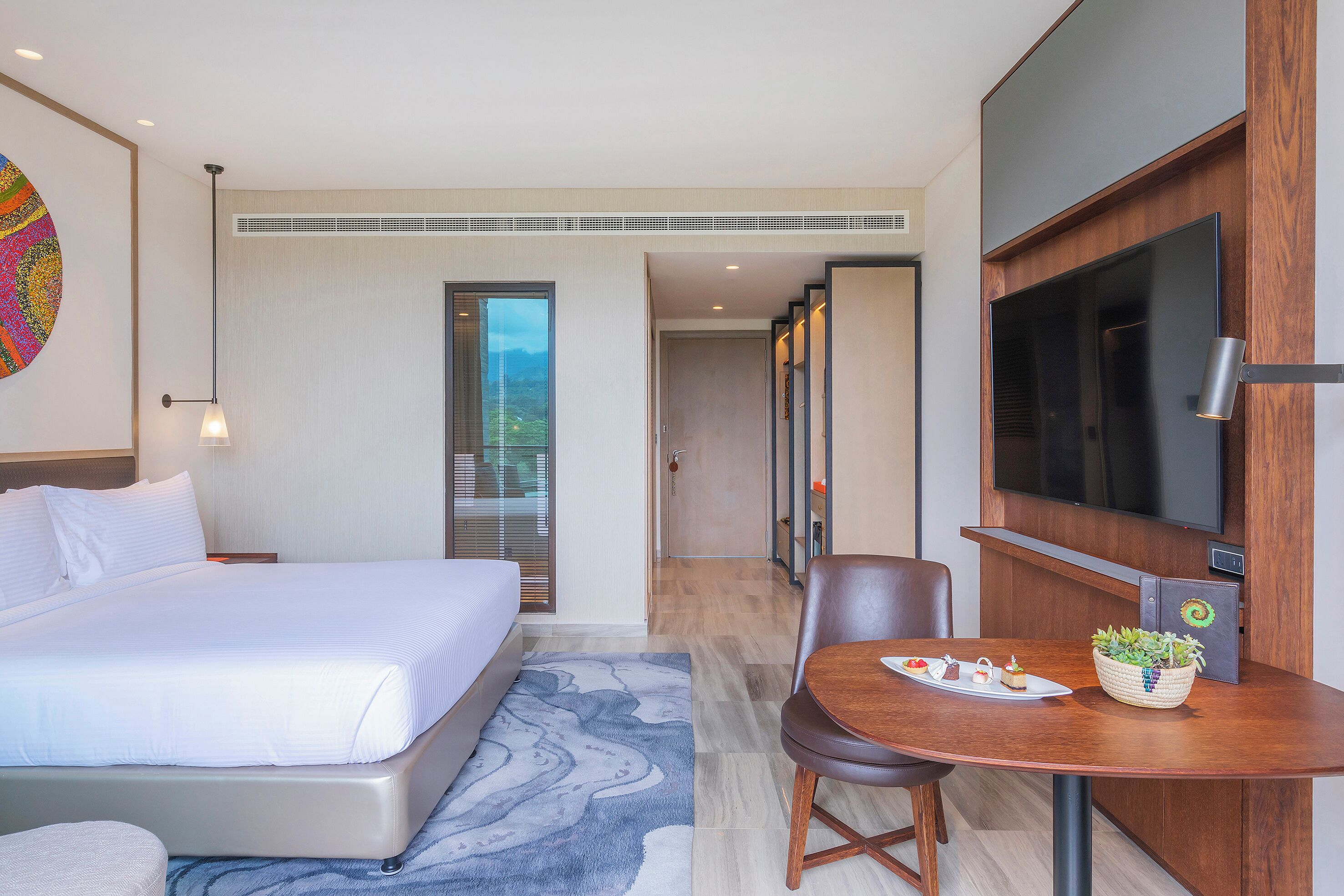 A hotel room with a bed, a desk with a plant, a TV, and a view of the outdoors.