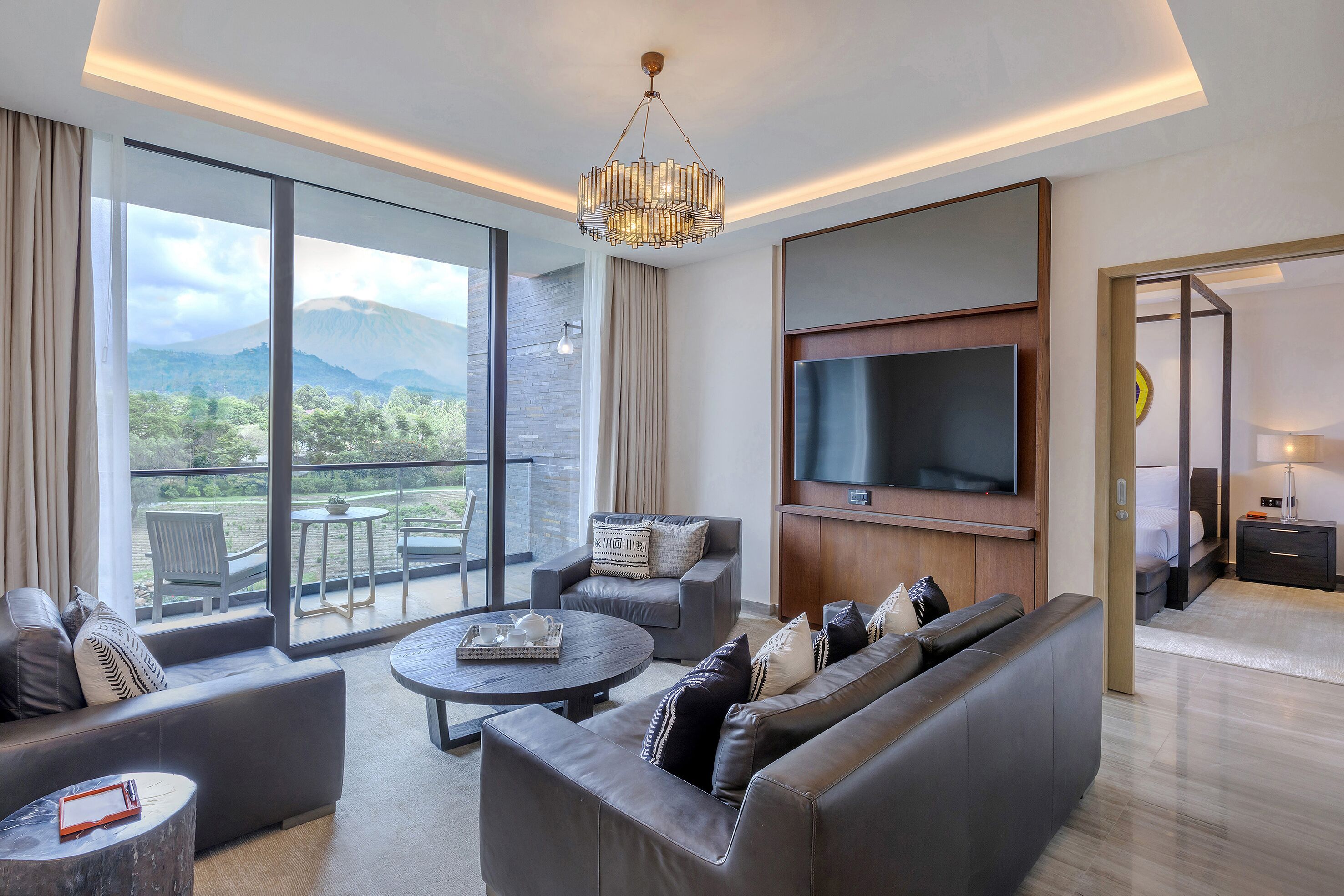 A modern hotel room with a large TV, comfortable seating, a balcony with a mountain view, and a stylish chandelier.