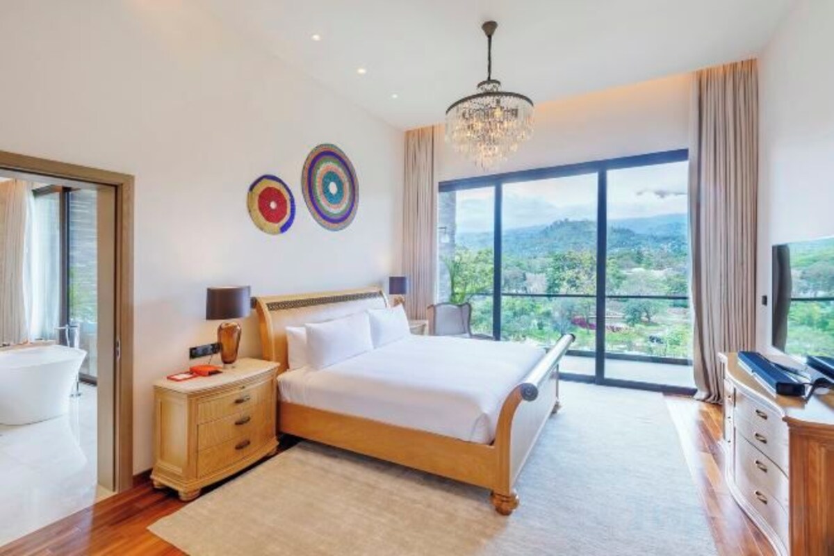 A spacious bedroom with a large bed, a nightstand, a dresser, and a television. There is a large window offering a view of the outdoors.