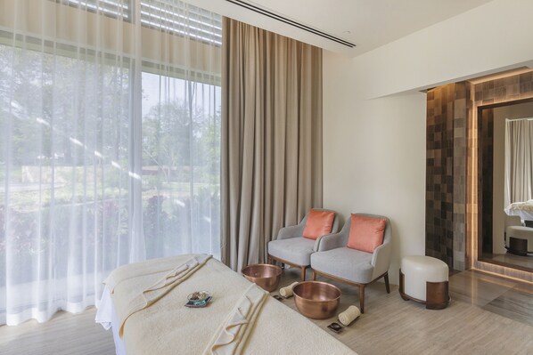 A modern hotel room with a large window, a sofa, a small table, and a massage table with towels and copper bowls. - Gran Melia Arusha (Arusha)