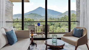 Habitación con un ventanal grande que ofrece vistas a una montaña y vegetación exuberante. Hay un sofá con un cojín azul, una silla y una pequeña mesa con un florero.