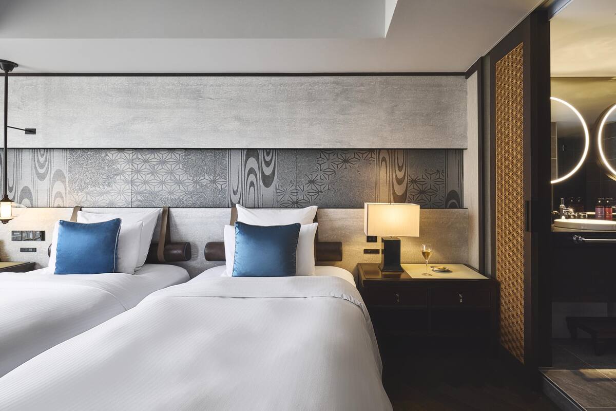 Ein Hotelzimmer mit zwei Betten, einem Nachttisch mit Lampe und einem rechts befindlichen Badezimmer.