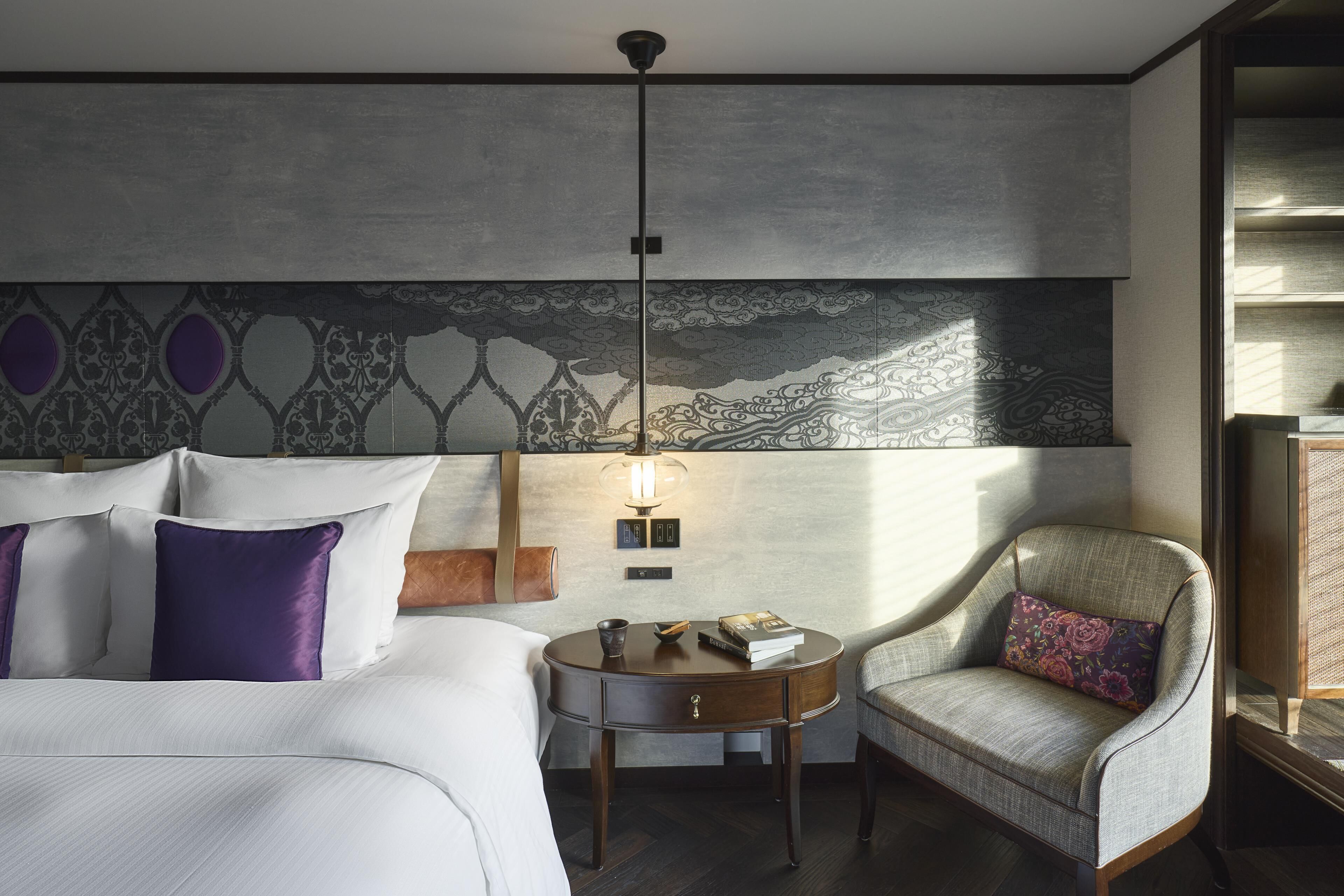 Una habitación de hotel con una cama, una mesita de noche redonda de madera, una silla y una pared con un diseño decorativo.