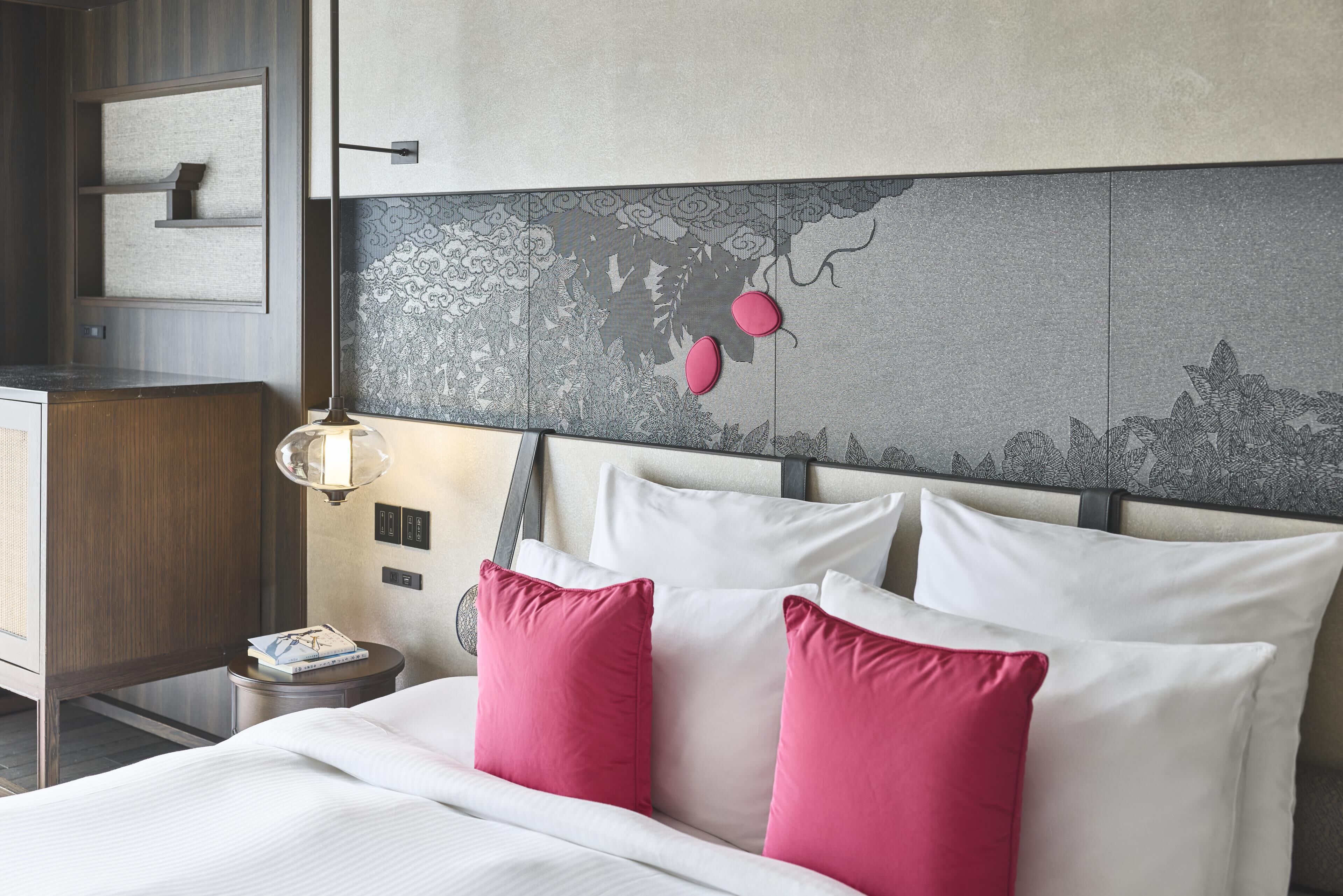 Habitació d'hotel amb llit, almoines, una tauleta de nit, làmpada i un mur amb un panell decoratiu.