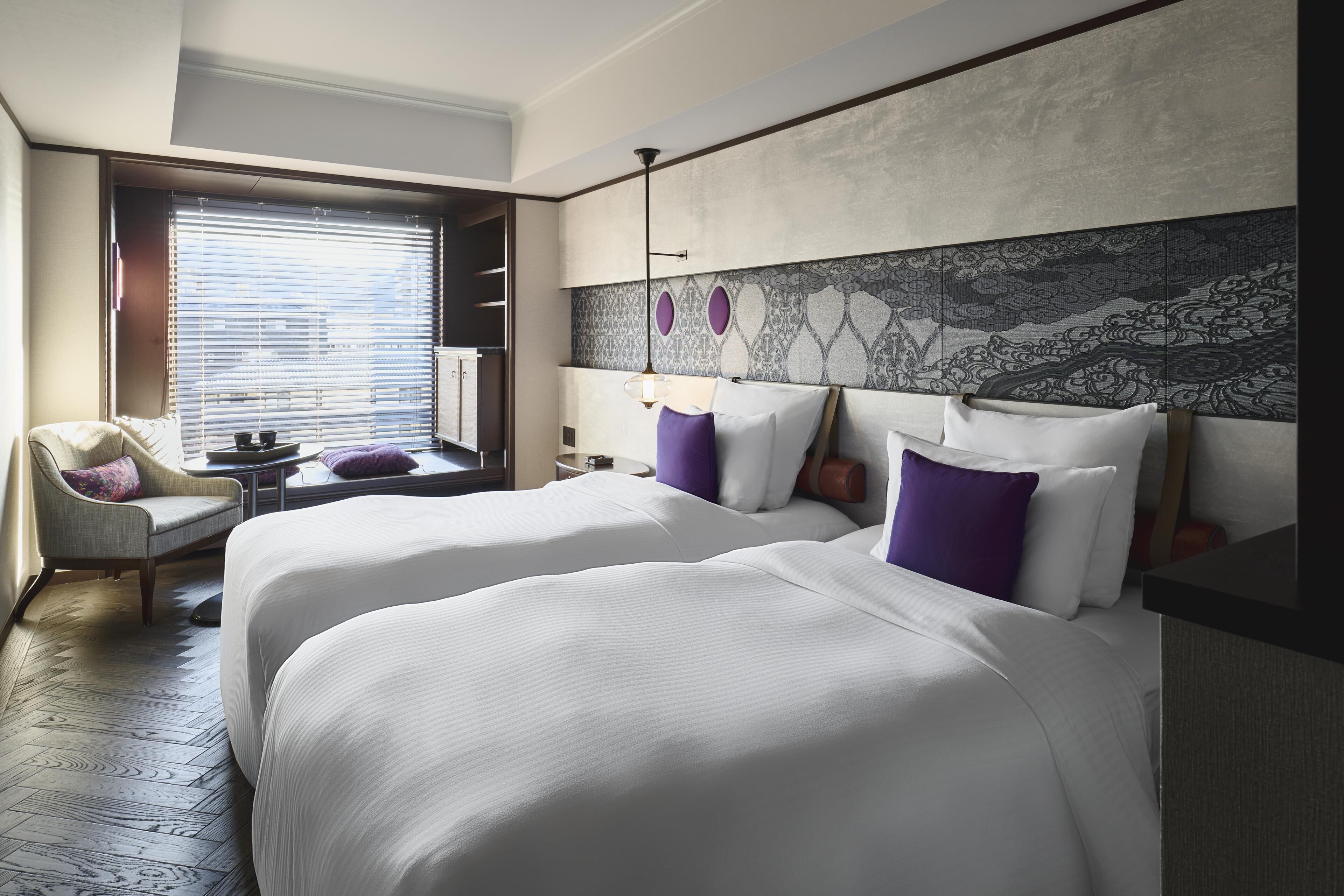 A hotel room with two beds, a chair, a window with blinds, and a decorative wall panel.