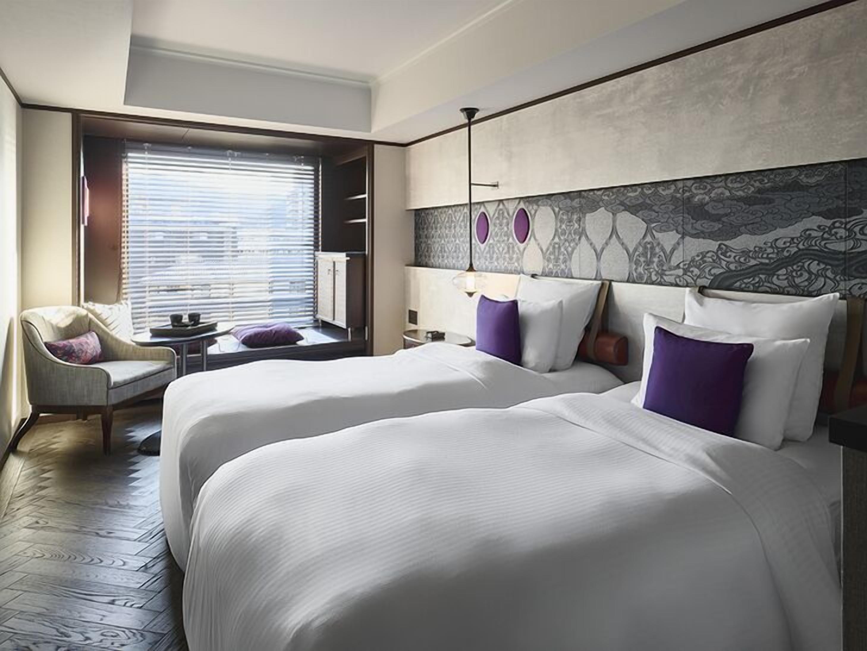 Een hotelkamer met twee bedden, een stoel, een raam met zonwering en decoratieve wand.