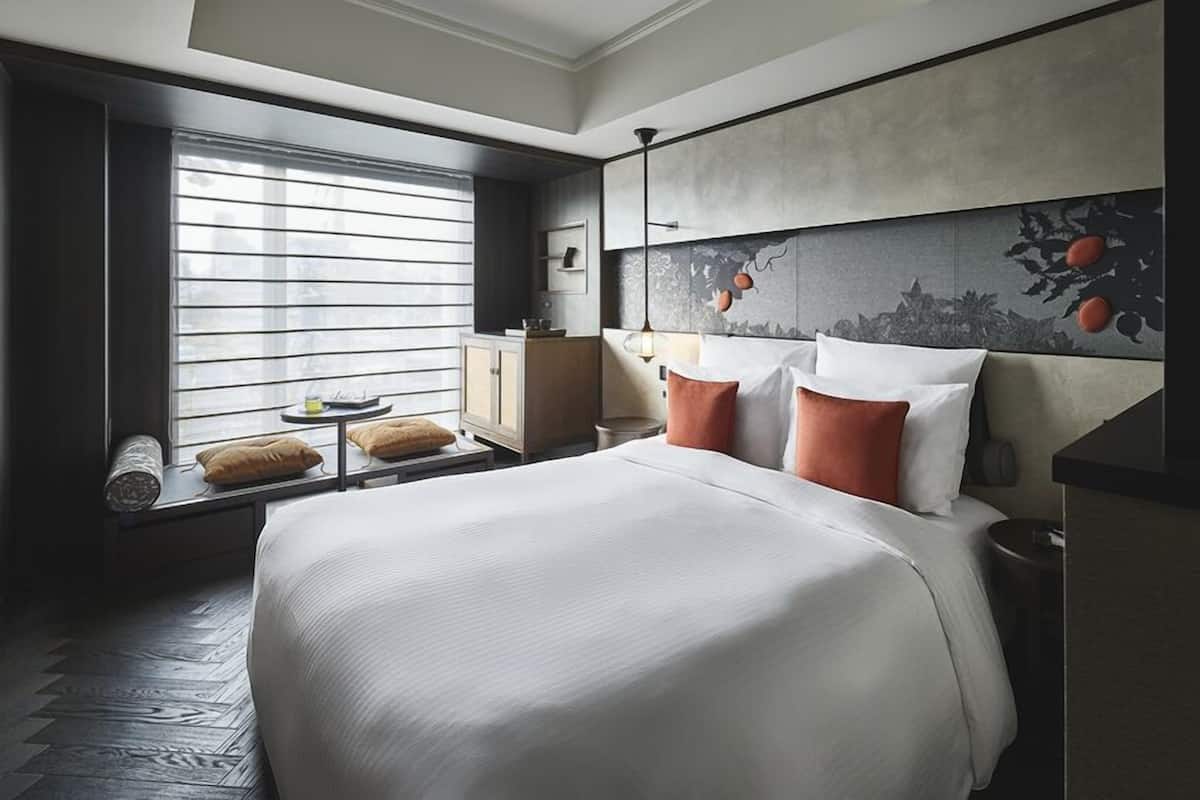 A hotel room with a large bed, a window with blinds, a nightstand, and a wall-mounted shelf.