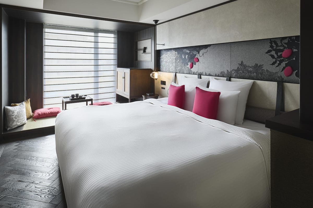Ein modernes Hotelzimmer mit einem großen Bett, einem Nachttisch, einem Schreibtisch und einem Fenster mit Jalousien.