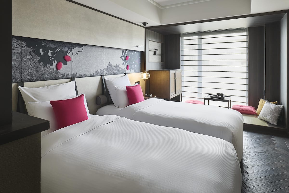 Ein Hotelzimmer mit zwei Betten, einem Fernseher, einem Fenster mit Jalousien und einem kleinen Tisch mit Lampe.