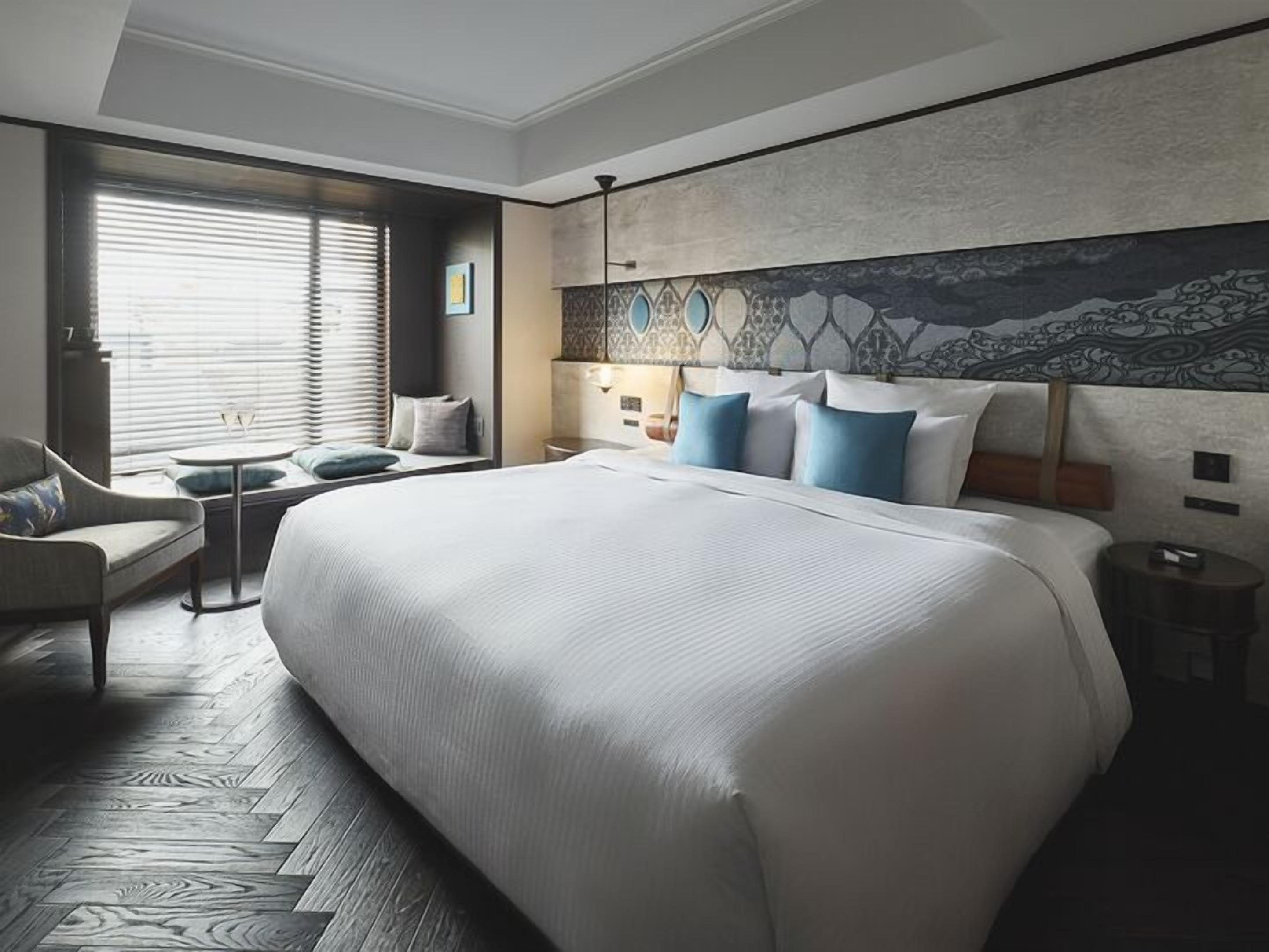 Een hotelkamer met een groot bed, een zithoek met een stoel en een klein tafeltje, een raam met zonwering, en een decoratieve wand.