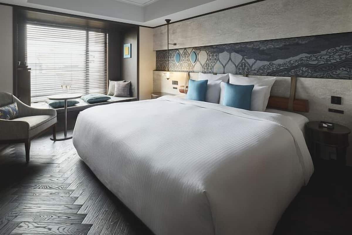 A hotel room with a large bed, a seating area with a chair and a small table, a window with blinds, and a decorative wall.