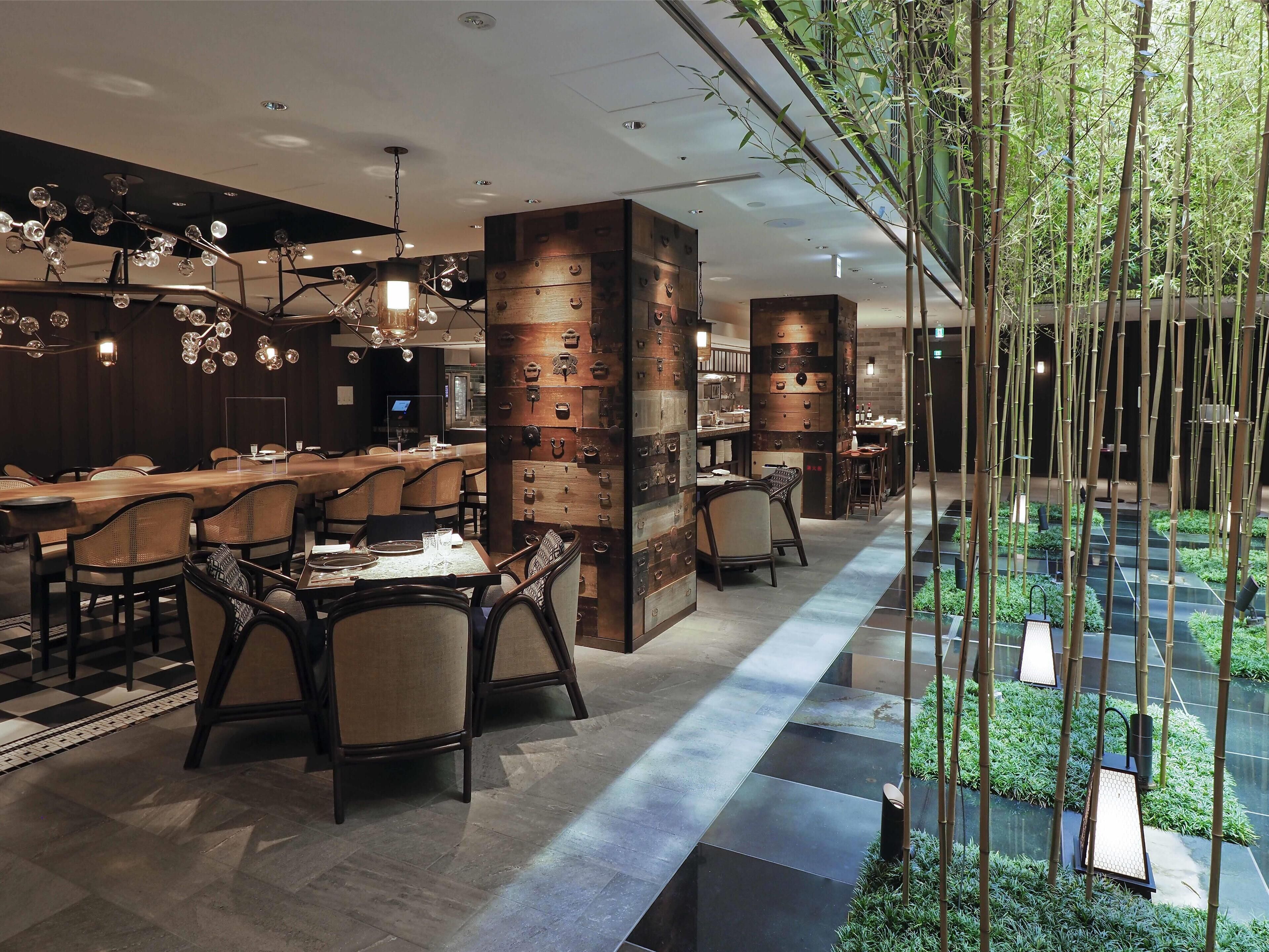 Un restaurante moderno con mesas y sillas de madera, una columna central con detalles decorativos y un área con jardín de bambú.