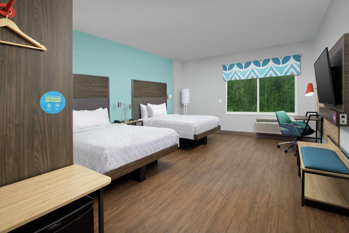 A hotel room with two beds, a desk, a chair, a TV, and a window with a view of greenery.