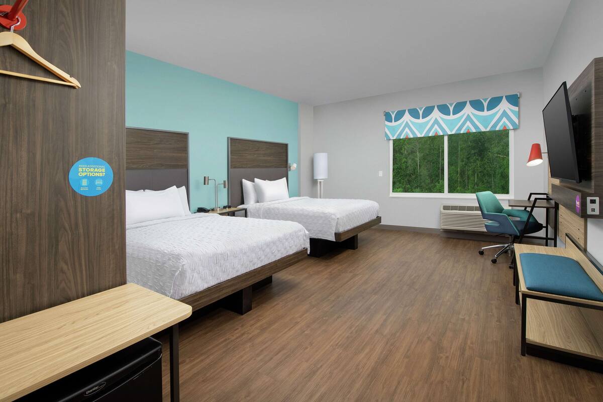 A hotel room with two beds, a desk, a chair, a TV, and a window with a view of greenery.