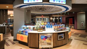 A modern hotel lobby with a central counter displaying snacks and beverages, two digital screens above the counter, and a seating area with a view of the lobby. - Tru by Hilton Lake City (Lake City)