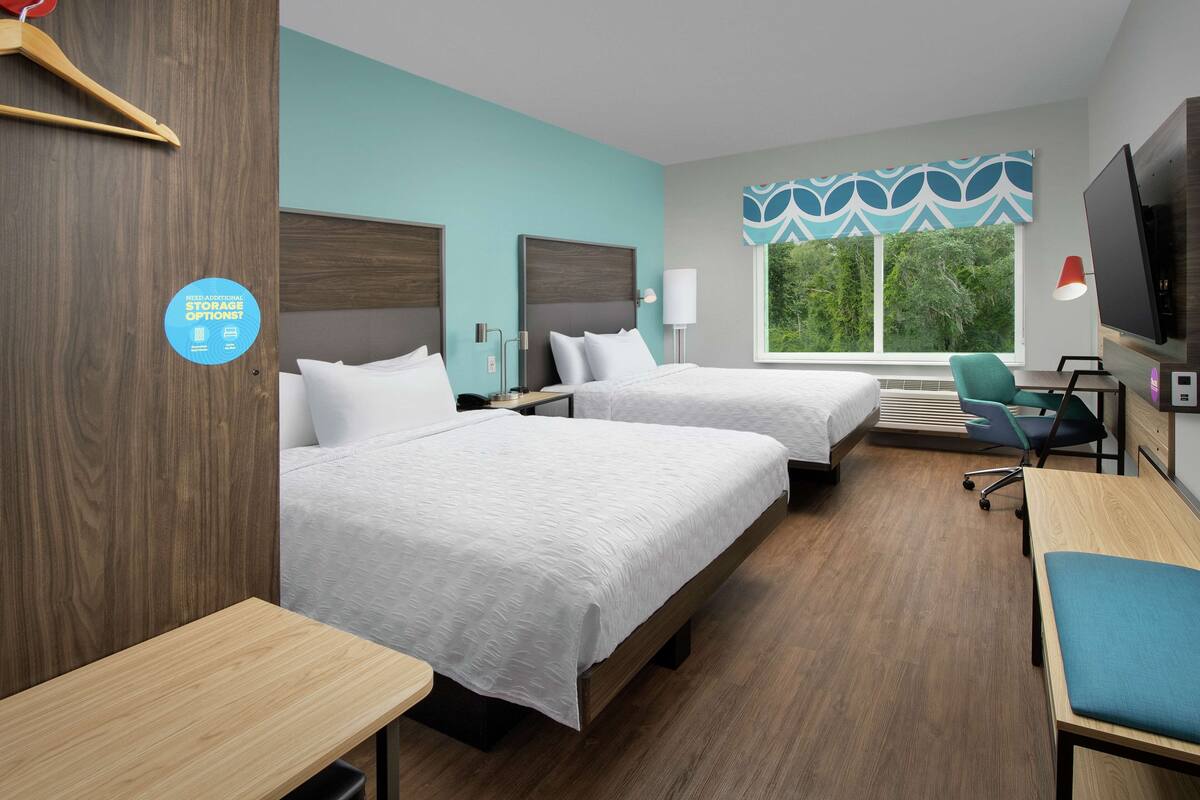 A hotel room with two beds, a desk, a chair, a TV, and a window with a view of greenery.