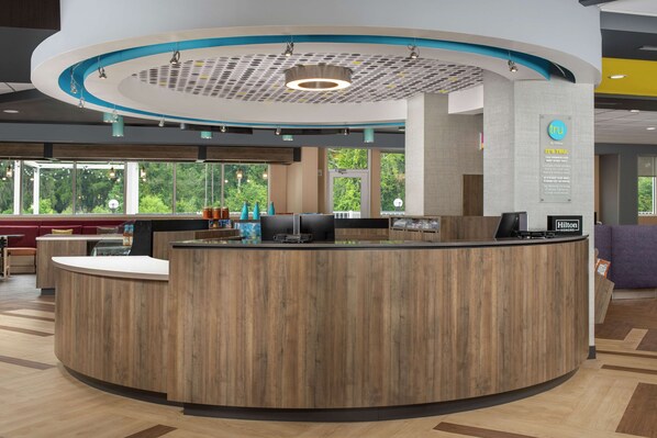Un área de recepción de hotel moderna, con un mostrador de madera, ventanas amplias que ofrecen vistas a la vegetación y un techo con un patrón geométrico único.