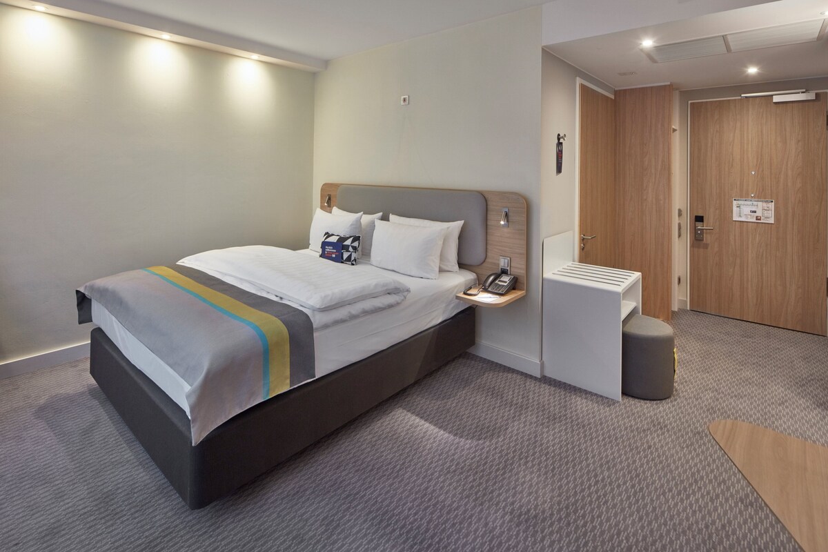 A modern hotel room with a large bed, bedside table, and a small seating area.