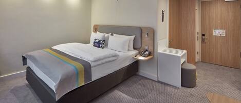 A modern hotel room with a large bed, bedside table, and a small seating area.
