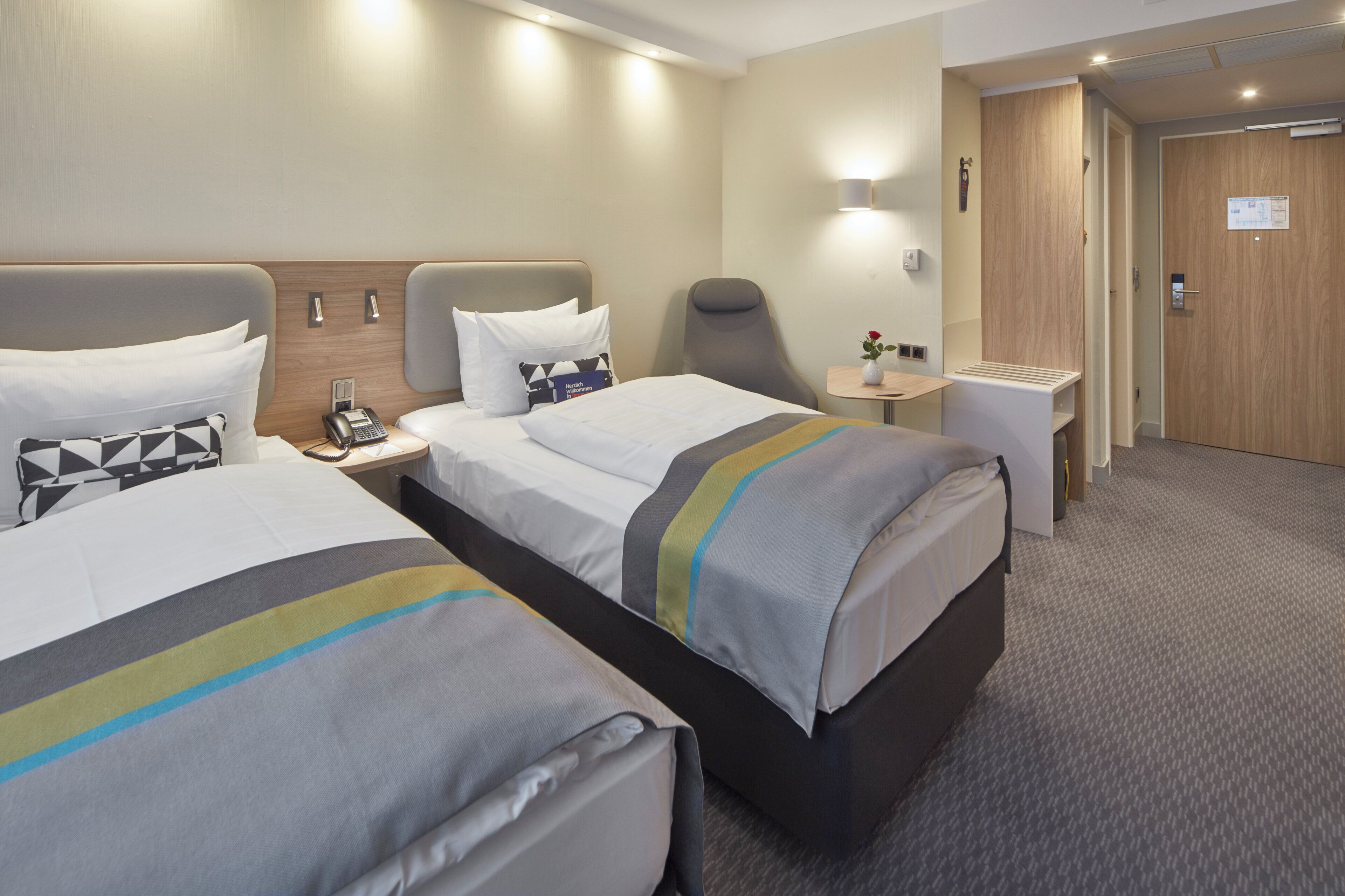A hotel room with two beds, a bedside table with a phone, a chair, and a small table with a vase.