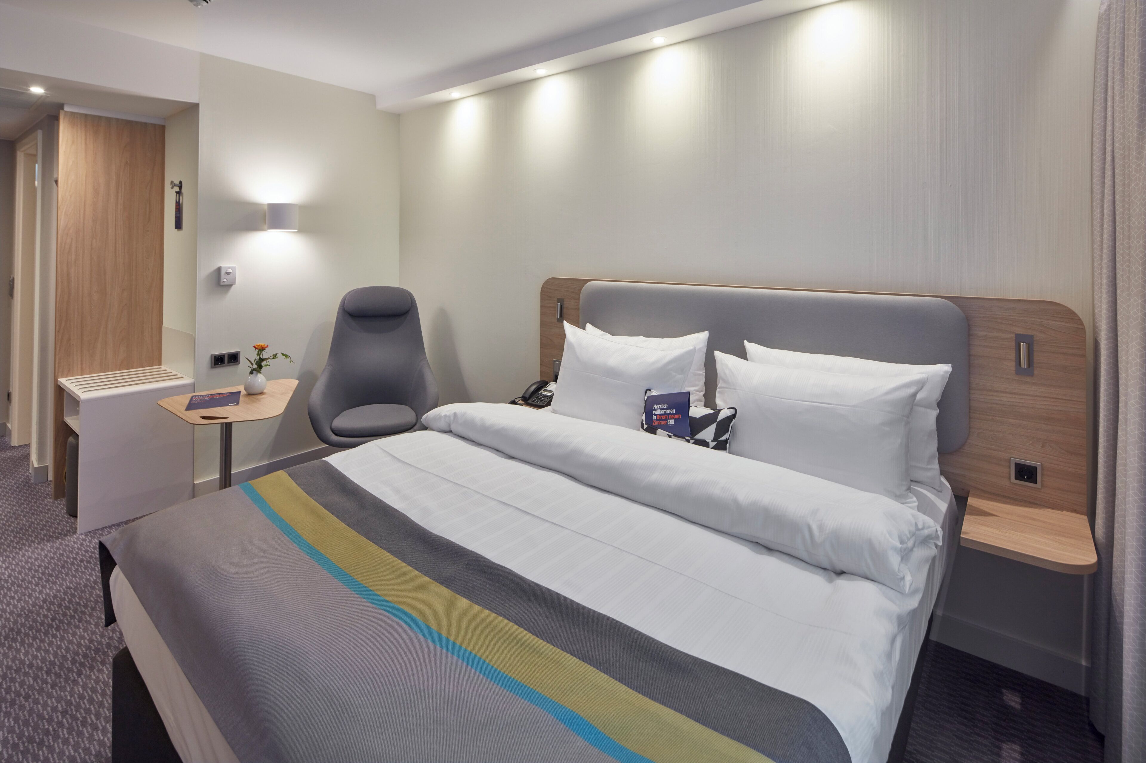 A hotel room with a bed, bedside table, chair, and a wall-mounted lamp.