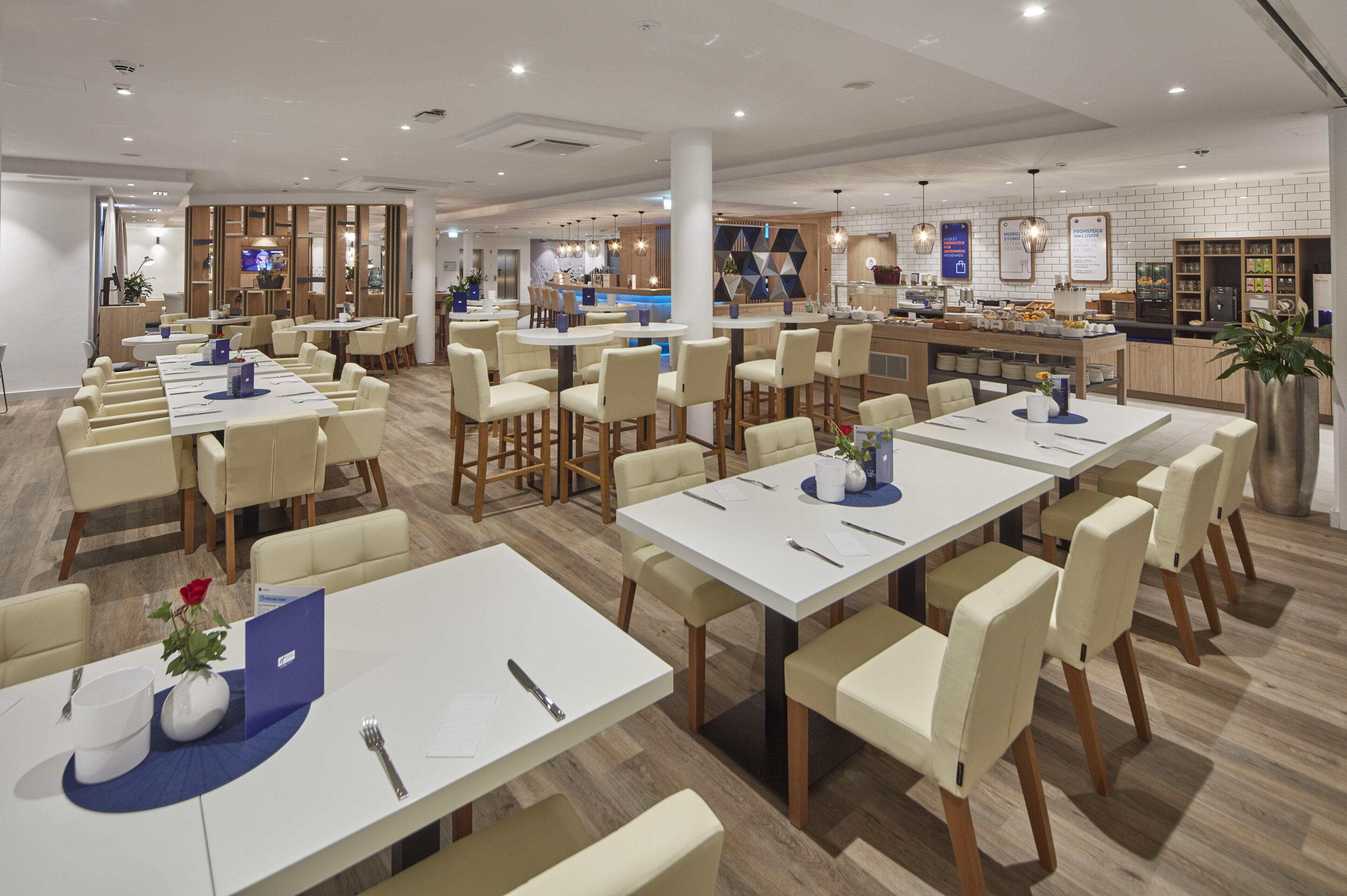 A modern dining area with multiple tables and chairs, a kitchen in the background, and a clean, well-lit environment.