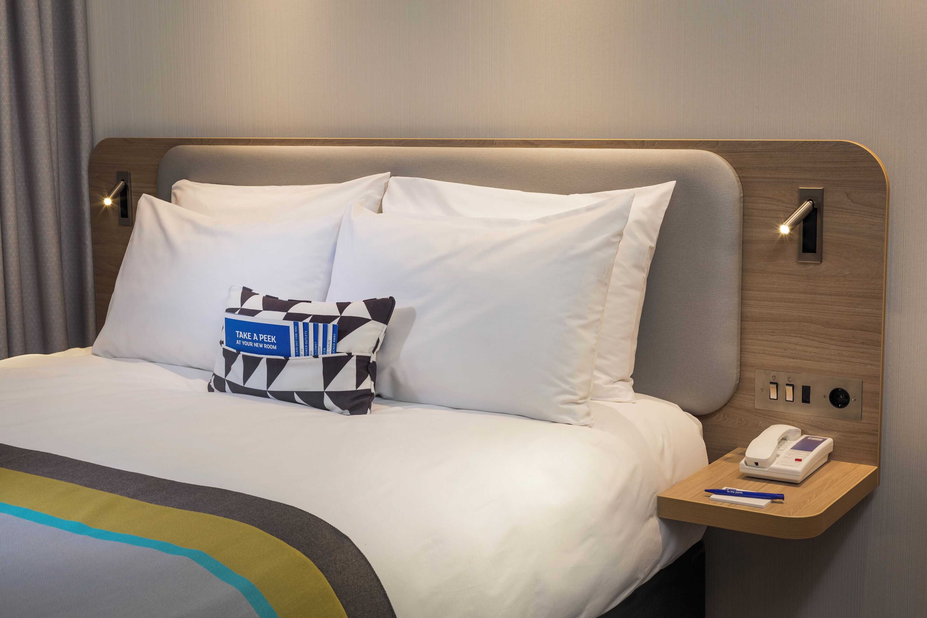 A neatly made bed with white linens, a bedside table with a phone and pen, and a pillow with a blue tag.