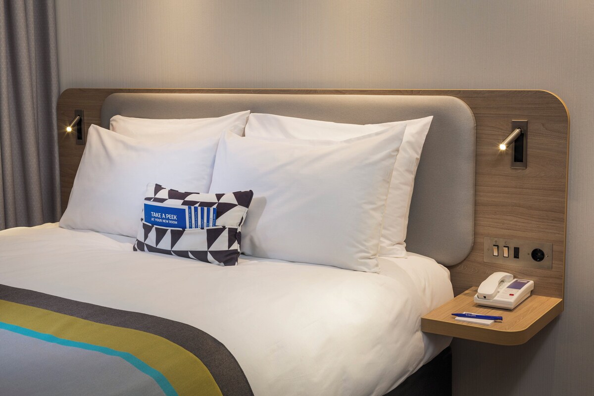 A neatly made bed with white linens, a bedside table with a phone and pen, and a pillow with a blue tag.